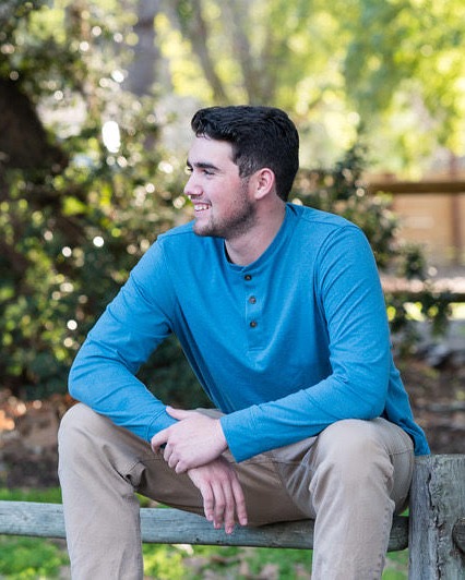 Samuel Fox, founder of Fox Marketing Systems, sitting on a wooden fence outdoors and smiling